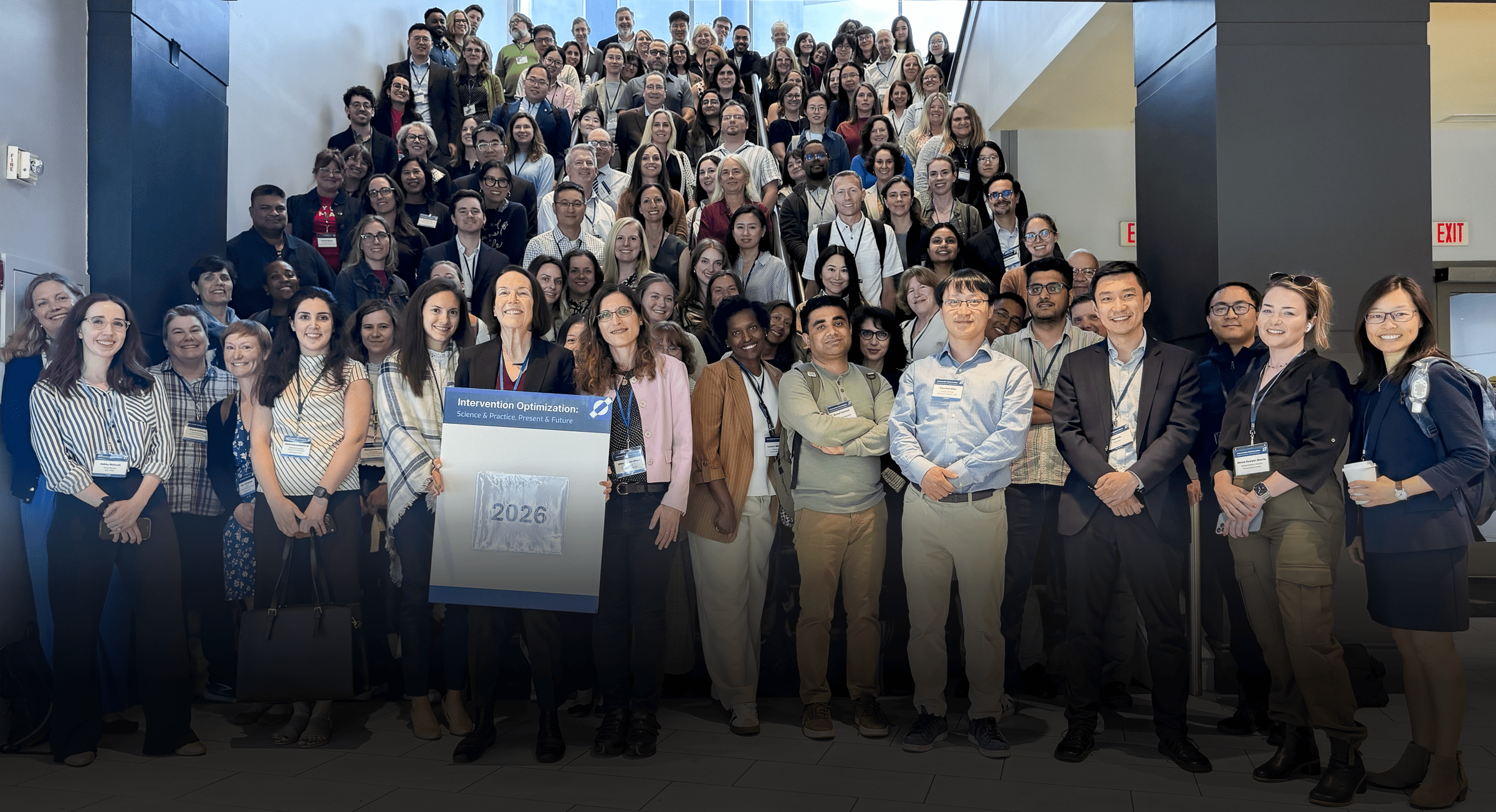 Group photo of roughly 150 conference attendees gathered on and around a staircase in an atrium-style lobby. Two people in the front row hold a sign reading "Intervention Optimization: Science & Practice, Present & Future" with the year "2026." Attendees wear name badges on lanyards and are dressed in business casual attire; exit signs and glass doors are visible in the background.