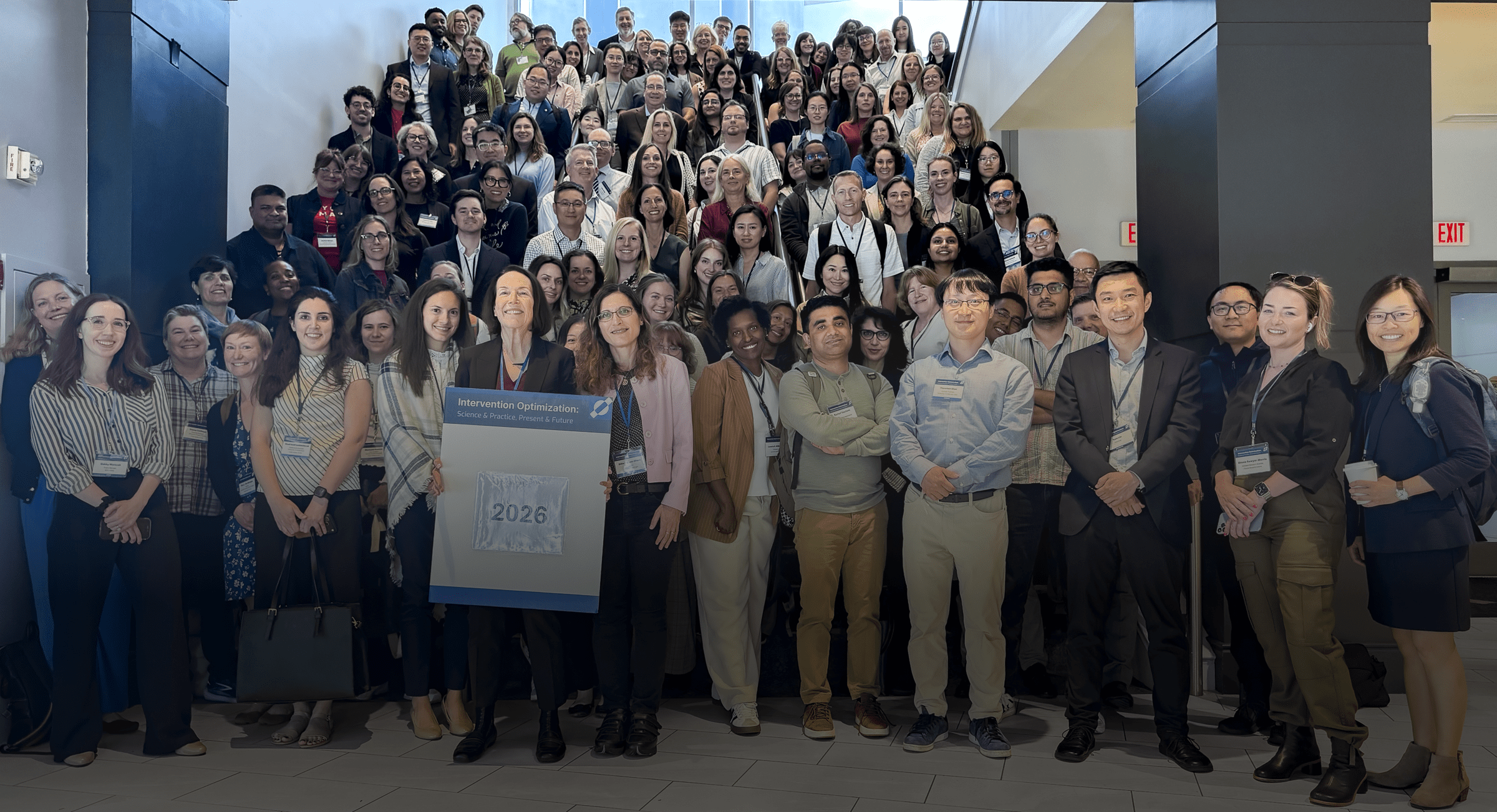 Group photo of roughly 150 conference attendees gathered on and around a staircase in an atrium-style lobby. Two people in the front row hold a sign reading "Intervention Optimization: Science & Practice, Present & Future" with the year "2026." Attendees wear name badges on lanyards and are dressed in business casual attire; exit signs and glass doors are visible in the background.